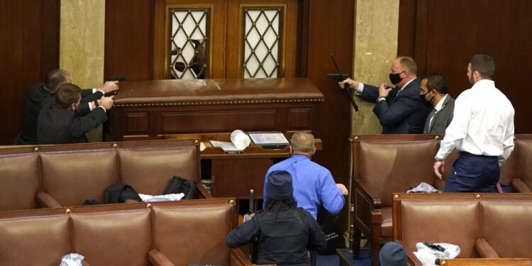El interior del recinto de la Cámara de Representantes con una ventana rota y un grupo de fuerzas de seguridad del Capitolio apuntando sus armas a un presunto manifestante